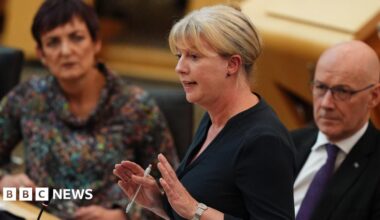 Shona Robison speaking in the Scottish Parliament. She has blonde hair and is wearing a black top, and is holding her hands out, holding a pen, while talking. Angela Constance is seated to her left and John Swinney is sitting to her right.