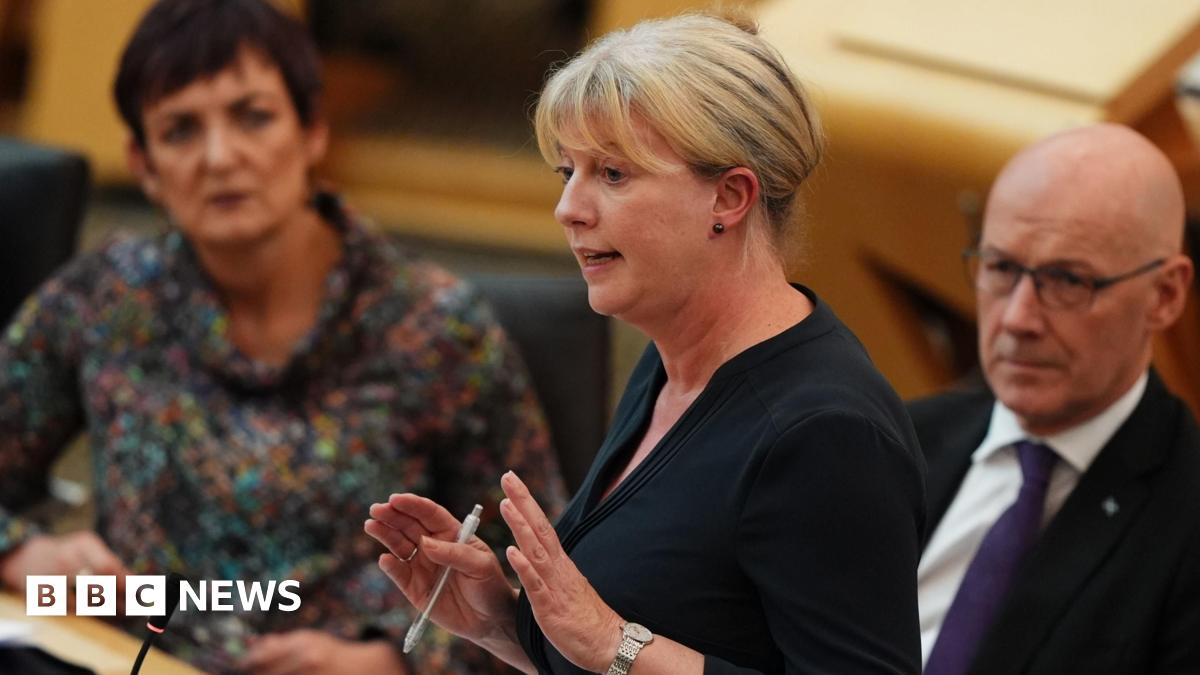 Shona Robison speaking in the Scottish Parliament. She has blonde hair and is wearing a black top, and is holding her hands out, holding a pen, while talking. Angela Constance is seated to her left and John Swinney is sitting to her right.