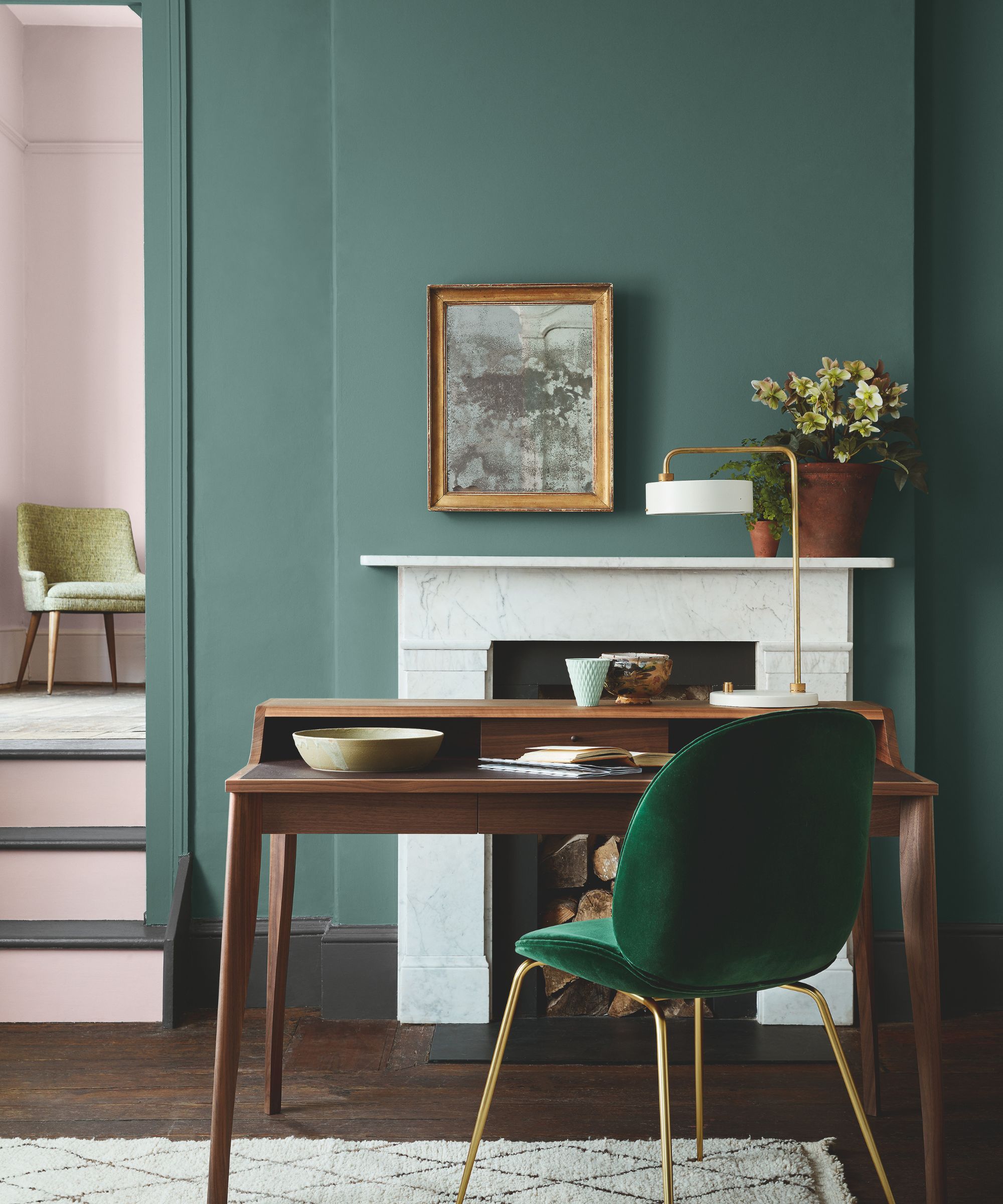 A home office with a teal wall, a white fireplace and a wooden desk with a green chair.