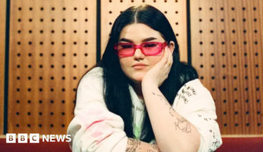 Mae Stephens has long dark hair, pink glasses, and sits in a recording studio, leaning forward with one arm resting on a table and the other propping up her face. She is wearing a light-coloured sweatshirt covered in handwritten-style illustrations and text, with several visible tattoos along her arms. Behind her is a wooden acoustic panel wall with evenly-spaced circular sound‑dampening holes, giving the space a warm, studio-like atmosphere.