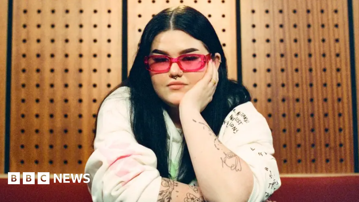 Mae Stephens has long dark hair, pink glasses, and sits in a recording studio, leaning forward with one arm resting on a table and the other propping up her face. She is wearing a light-coloured sweatshirt covered in handwritten-style illustrations and text, with several visible tattoos along her arms. Behind her is a wooden acoustic panel wall with evenly-spaced circular sound‑dampening holes, giving the space a warm, studio-like atmosphere.