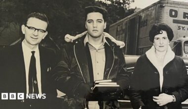 A black and white photograph of three people. The man in the centre is Elvis Presley wearing a shirt, a black, fleeced leather jacket and black gloves. He is holding three books. The man to the left has black hair, black rimmed glasses, a white shirt and a black tie. The woman on the right has dark short hair and a dark fleeced coat. Behind them is a van, a car and trees.