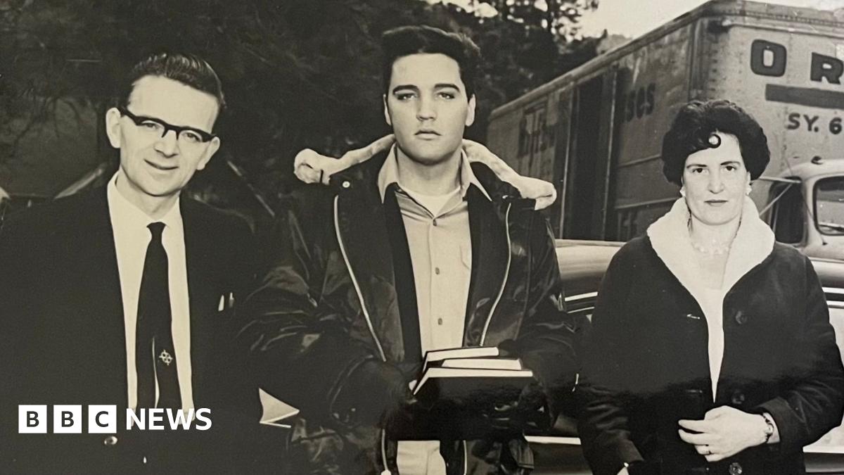 A black and white photograph of three people. The man in the centre is Elvis Presley wearing a shirt, a black, fleeced leather jacket and black gloves. He is holding three books. The man to the left has black hair, black rimmed glasses, a white shirt and a black tie. The woman on the right has dark short hair and a dark fleeced coat. Behind them is a van, a car and trees.
