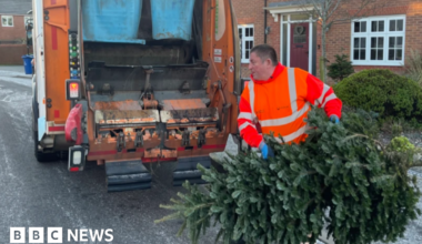 A man in a bright hi-viz jacket is lifting a large Christmas tree into the back of a wagon. The vehicle is parked outside houses