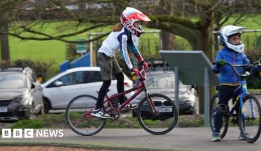 Child on a bike in a red helmet with red gloves and another child in a white helmet wearing blue. The background is a park like enviroment.