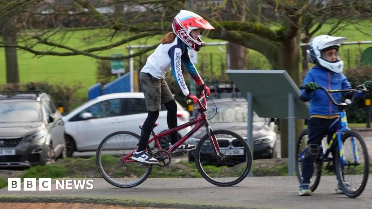 Child on a bike in a red helmet with red gloves and another child in a white helmet wearing blue. The background is a park like enviroment.