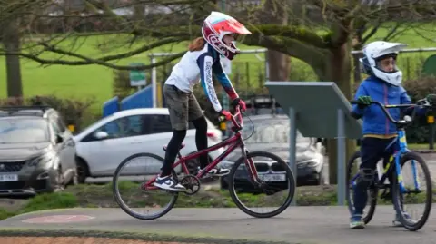 BBC Child on a bike in a red helmet with red gloves and another child in a white helmet wearing blue. The background is a park like enviroment.