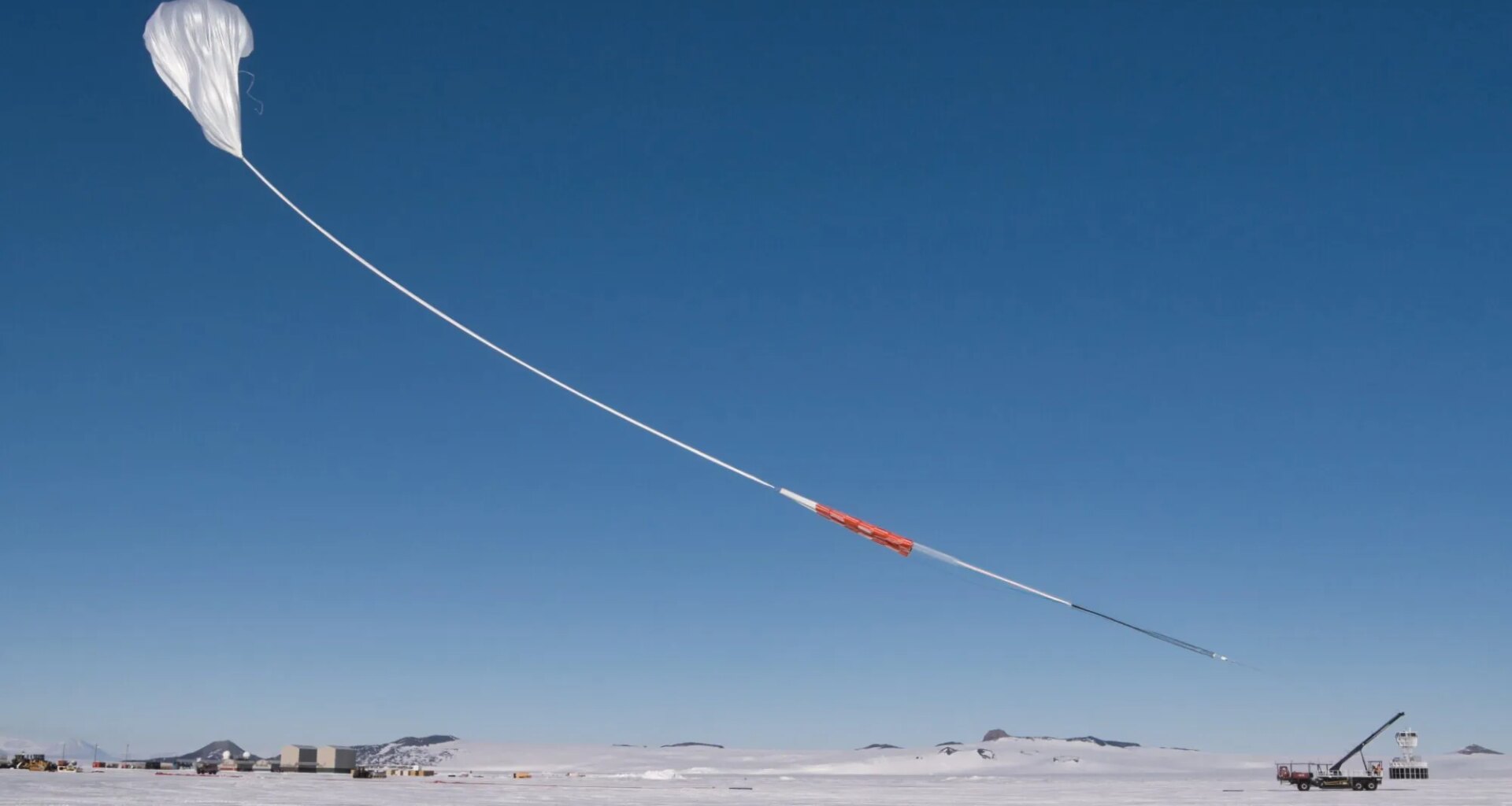 A white scientific balloon rises into the bright blue sky above snowy Antarctica. The sky and the balloon take up most of the image, with the ground only being a small white stripe at the bottom of the photo. The balloon has a long white "tail" with orange and black portions.