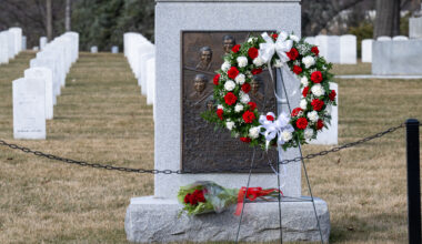 A wreath with red and white flowers, green leaves, and white ribbon bows rests on a tripod stand with three thin metal legs. It stands in front of the Space Shuttle Challenger Memorial. The Memorial has two parts: A polished gray stone headstone with a bronze embossed plaque on it, resting on a small platform of rough gray stone. The plaque has the likeness of Challenger and the crew on it. Behind the memorial, we can see rows of white stone headstones.