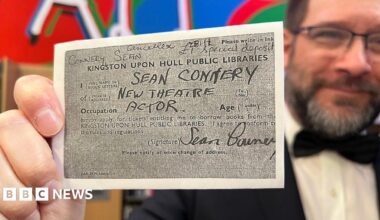 Librarian Matt Soare holds the photocopied card close to the camera. He has dark hair and beard and is wearing glasses and a dinner jacket and black bow tie