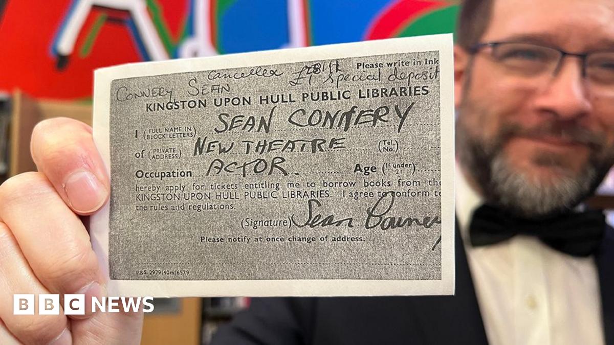 Librarian Matt Soare holds the photocopied card close to the camera. He has dark hair and beard and is wearing glasses and a dinner jacket and black bow tie