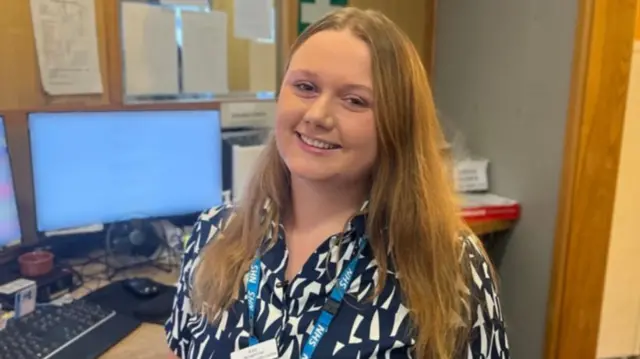 A woman with long dark blonde hair is sitting in front of a computer screen with a blue NHS lanyard round her neck