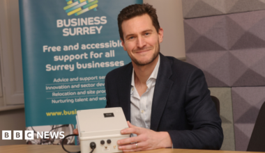 The image shows a man smiling for a photograph, while holding a white electrical object. He is wearing a navy blue suit with a striped undershirt. There is a blue banner behind him that reads: "Business Surrey, Free and accessible support for all Surrey businesses" with some smaller text underneath.