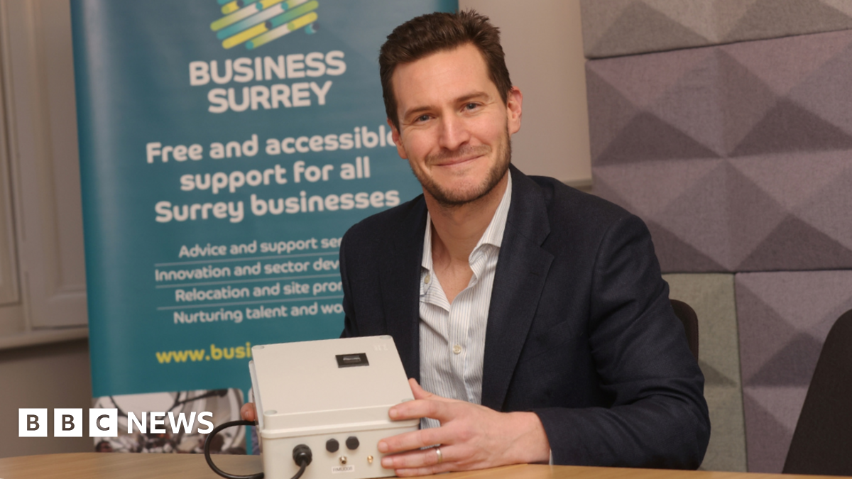 The image shows a man smiling for a photograph, while holding a white electrical object. He is wearing a navy blue suit with a striped undershirt. There is a blue banner behind him that reads: "Business Surrey, Free and accessible support for all Surrey businesses" with some smaller text underneath.