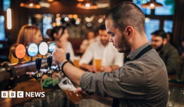 A bartender, with short black hair and a black stubble, wearing a grey shirt, standing behind a bar. He is pulling a pint of beer. There are four people in the background, which is blurred.
