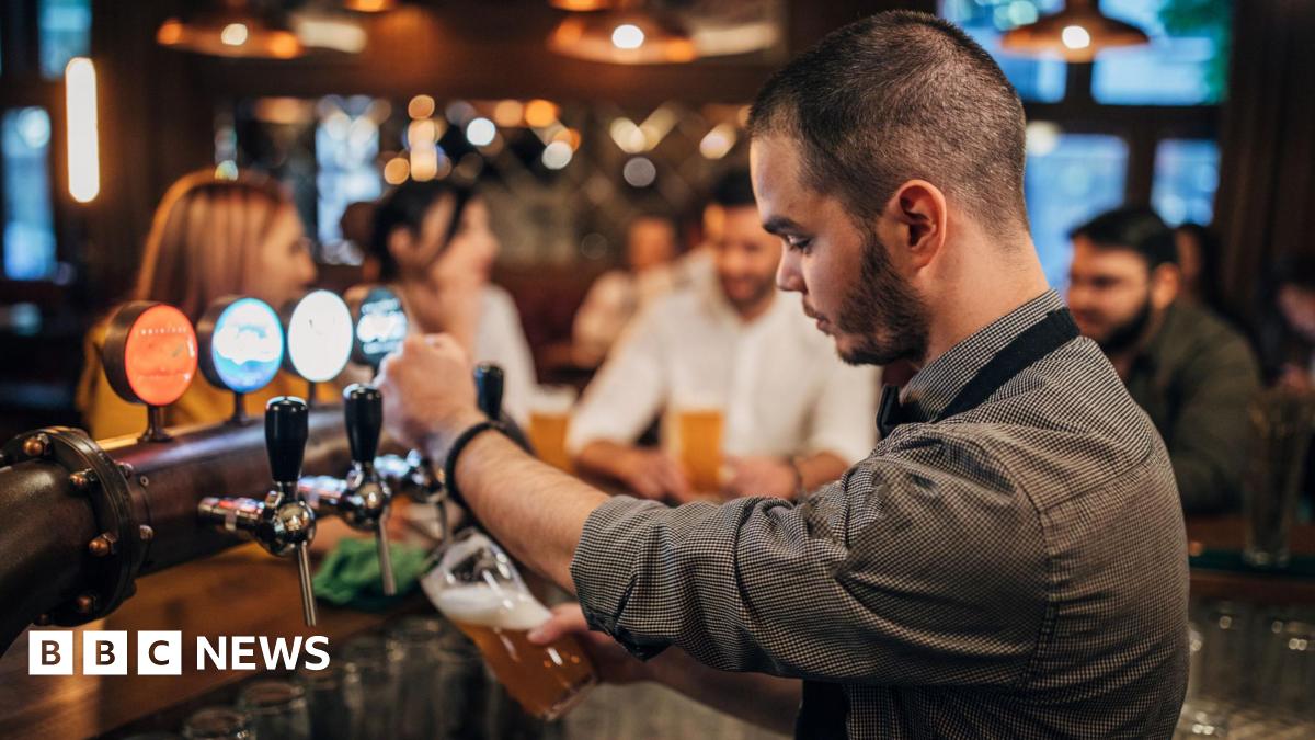 A bartender, with short black hair and a black stubble, wearing a grey shirt, standing behind a bar. He is pulling a pint of beer. There are four people in the background, which is blurred.