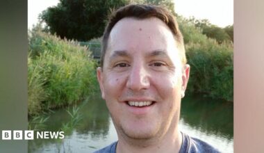 James Clements is standing in front of a small lake which is surrounded by tall grass. He has short brown hair, and is smiling at the camera. He is wearing a blue T-shirt.
