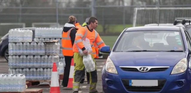 A man holding bottled water in a orange hi-vis jacket speaks to a person driving a blue car at a bottled water station