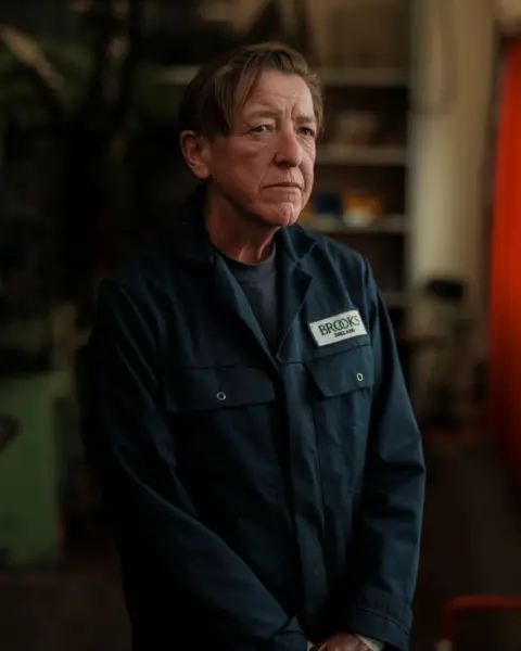 Portrait of Britain/Lloyd Wright Lisa looks ahead, side-on to the camera. She has short dark hair and is wearing a blue top and blue overalls, bearing the name Brooks. 