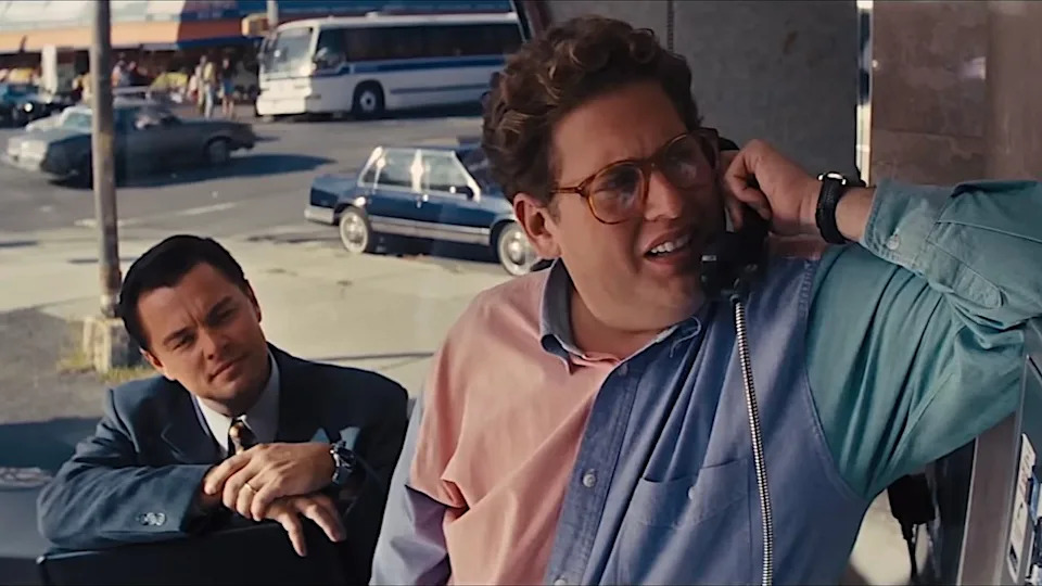 Jonah Hill talking on the phone with Leonardo DiCaprio holding a briefcase in the background during a scene in The Wolf of Wall Street.