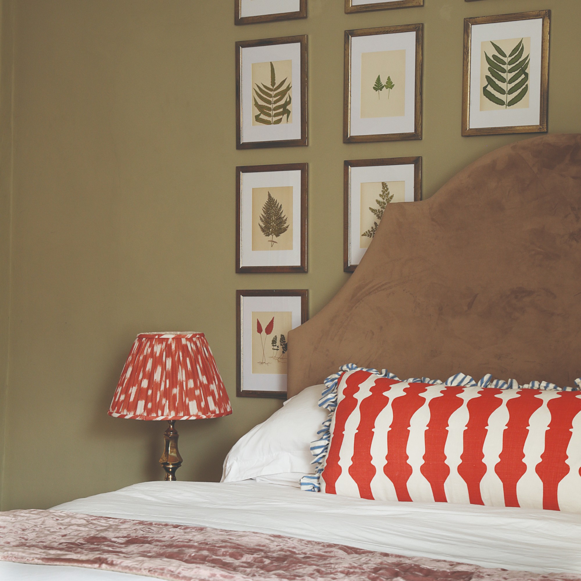 An olive green-painted bedroom with a brown fabric headboard, a leaf gallery wall and a table lamp with a patterned red shade
