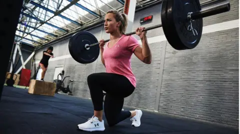 @vicswilliamson Vicky is kneeling down on one leg, in a gym, and holding a weight across her shoulders