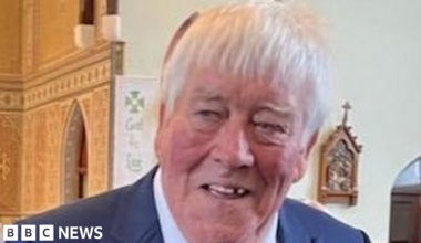 An elderly man with short white hair, a blue jacket and white shirt, smiles while he stands in a church.