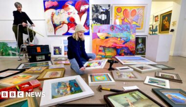 A woman sits on the floor of an art gallery, surrounded by paintings and artwork. Another woman is standing on a small stepladder in the background