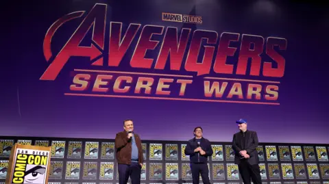 Getty Images Marvel bosses stand on stage at San Diego's Comic Con in 2024, when their rebooted plans for Doomsday and Secret Wars were announced. A large, red Avengers: Secret Wars logo is displayed on screen, with three men stood in front of it talking to an audience.