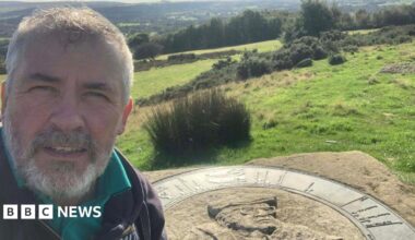 Nick Burton stands at the memorial to the late fell walker and writer Alfred Wainwright. The piece of stone has a picture of Wainwright carved into it. Nick stands on the left, he has short grey hair, a grey beard and wears a black coat with a teal polo shirt under it.
