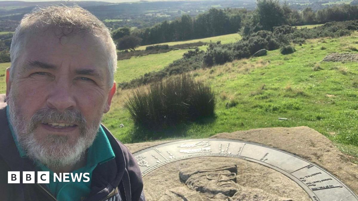 Nick Burton stands at the memorial to the late fell walker and writer Alfred Wainwright. The piece of stone has a picture of Wainwright carved into it. Nick stands on the left, he has short grey hair, a grey beard and wears a black coat with a teal polo shirt under it.