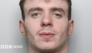A mugshot of a 20-year-old man. He has dark brown hair which is short on the sides and curly on the top, bushy eyebrows and minimal facial hair. He appears to be smirking slightly. He is wearing a grey sweatshirt and is standing in front of a pale, plain background.
