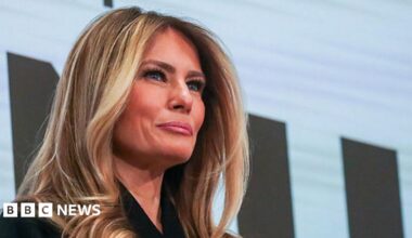 A head and shoulders shot of Melania Trump seen from beneath her as she stands on a balcony. The word 'film' can be seen behind her.
