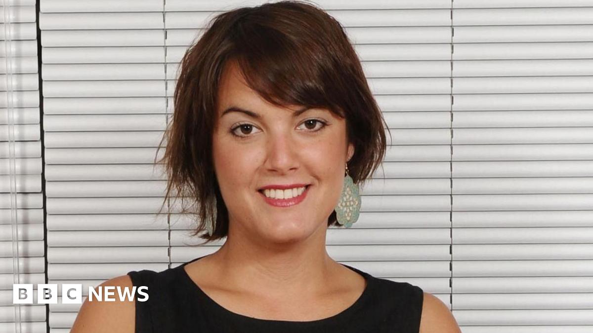 Yasmina Siadatan is sat on a sofa in front of closed white blinds. She has brown hair and is wearing green earrings and smiling for the picture.