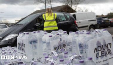 An image of packs of bottled water stacked up. Behind the bottles is a person with long blonde hair in a high-vis vest talking to someone through the window of a black car