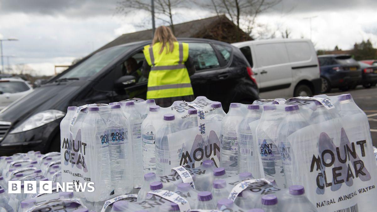 An image of packs of bottled water stacked up. Behind the bottles is a person with long blonde hair in a high-vis vest talking to someone through the window of a black car