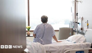 A man sits on a hospital bed facing a window. His back is facing the camera.