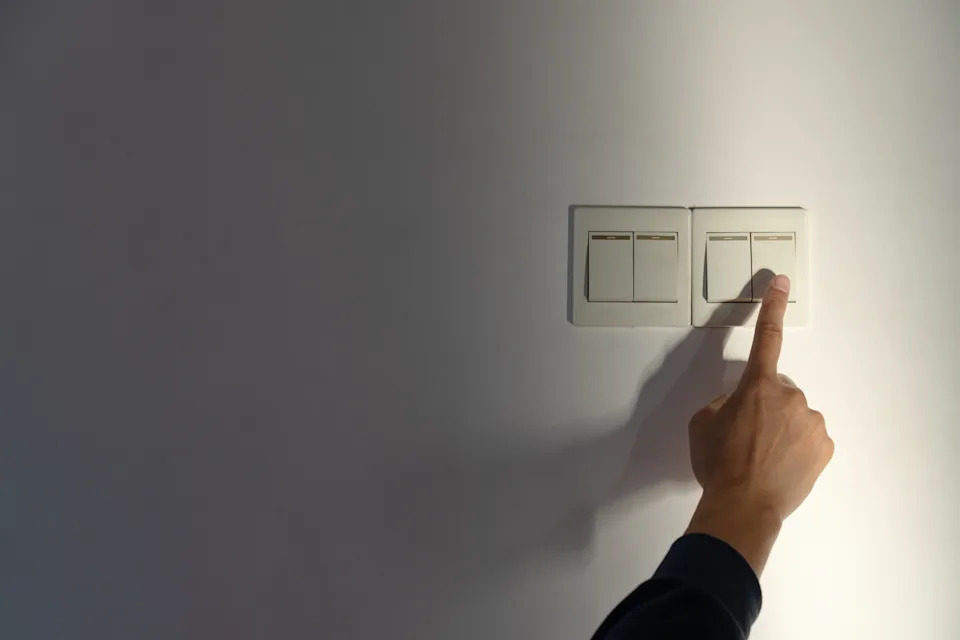 A close-up shot captures an individual's hand pressing a light switch on a plain wall. The scene is simple yet functional, highlighting the action of turning off or on a light source. The visual style is straightforward and utilitarian, emphasizing practicality over aesthetics. This image could be used for instructional materials, home improvement guides, or any context requiring a clear depiction of basic household tasks.