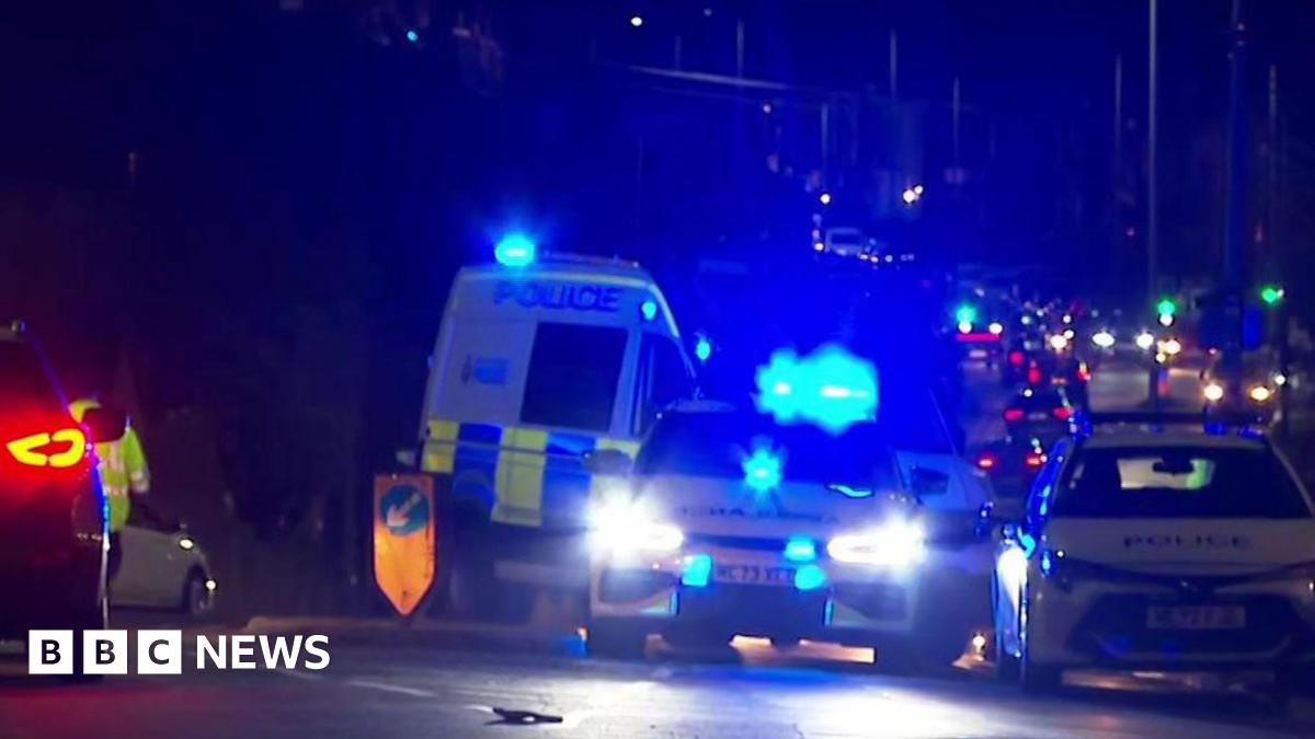 Emergency service vehicles including a police van and cars on Bolton Road in Ashton-in -Makerfield in front of a line of traffic. It is dark.
