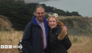 Terry, on the left, is wearing a patterned shirt and a black coat and has short brown hair. Wendy J. Tomlin-Hess is on the right and has short dark blonde hair, and is wearing a black coat. They are stood on a hill.