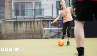 A woman with white blonde hair in a ponytail and a peach T-shirt with black leggings is kicking a ball on an outdoor football pitch. Someone blurred in the foreground is standing in front of her.