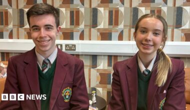 Two students, a boy and a girl, sitting side by side. The boy, with short brown hair, is sitting on the left side of the image and the girl, with blonde hair, tied up, is sitting on the right. They are wearing school uniforms, white shirts, dark green jumpers and burgundy blazers.