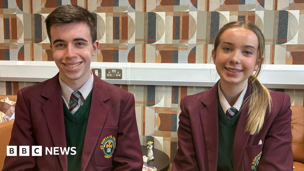 Two students, a boy and a girl, sitting side by side. The boy, with short brown hair, is sitting on the left side of the image and the girl, with blonde hair, tied up, is sitting on the right. They are wearing school uniforms, white shirts, dark green jumpers and burgundy blazers.
