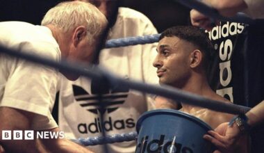 A boxer sits in the corner of a boxing ring, being attended to by his team - one holding an Adidas-branded bucket and others dressed in Adidas gear - offering support and guidance between rounds.
