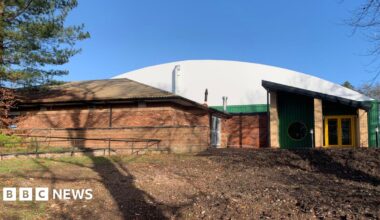 An indoor tennis centre building with a curved white roof and green and yellow entrance doorway. There is another low, brick building in front of it and rough ground in front of that. Shadows of trees are cast across the buildings.