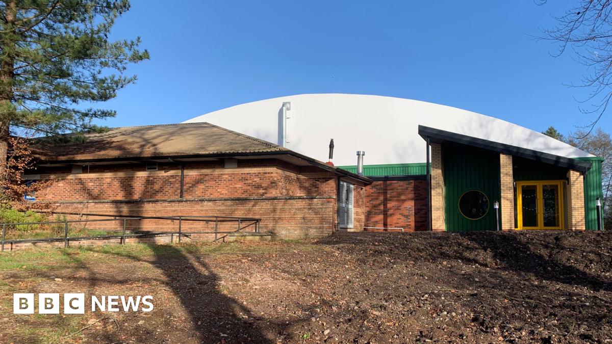 An indoor tennis centre building with a curved white roof and green and yellow entrance doorway. There is another low, brick building in front of it and rough ground in front of that. Shadows of trees are cast across the buildings.