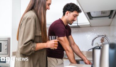 Couple stand at a kitchen sink with the man's hands in the sink with the tap running and a woman next to him holding a glass
