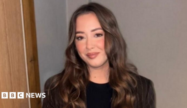 Laura is a young woman with long, brown hair. She is smiling at the camera. She is standing against a white wall and is wearing a brown pinstripe blazer and a black t-shirt.