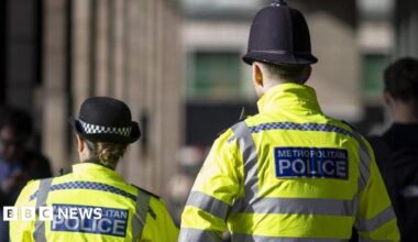 Two police officers, one shorter woman on the left and a tall man on the right, walk with backs to the camera. They wear fluorescent jackets which have a blue patch on them which reads 'Metropolitan Police'. They also have black hats.