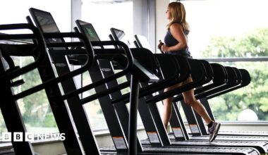 A stock image of a woman running on a treadmill. She is in a gym with floor to ceiling windows in front of her and next to her. She is running on one of six treadmills lined up and the others are empty.